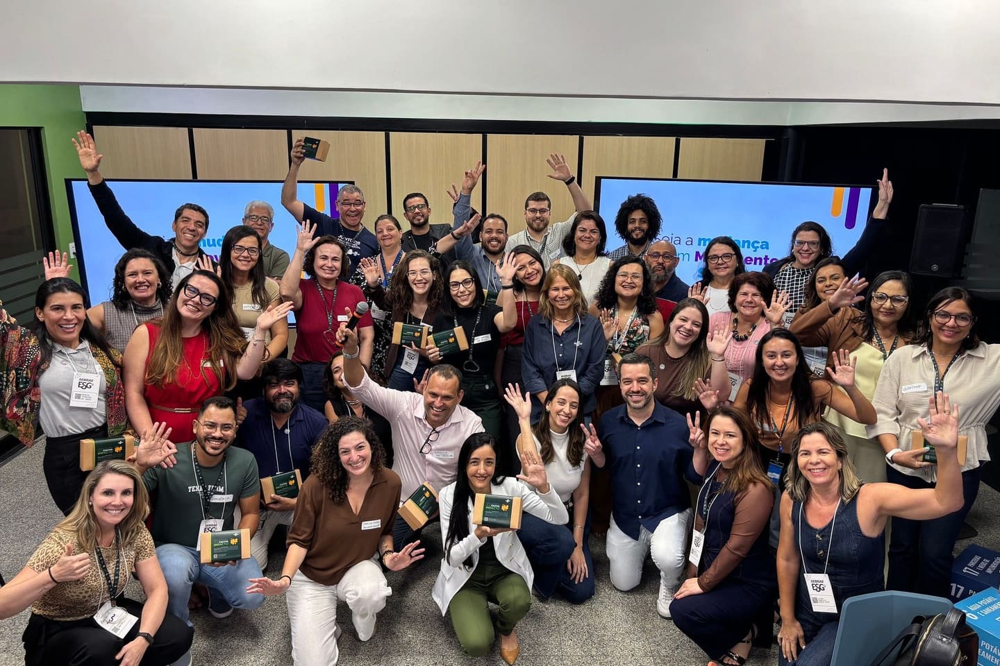 Turma de cerca de quarenta participantes com as mãos levantadas ao fim da oficina, diante das telas do Movimento Circular