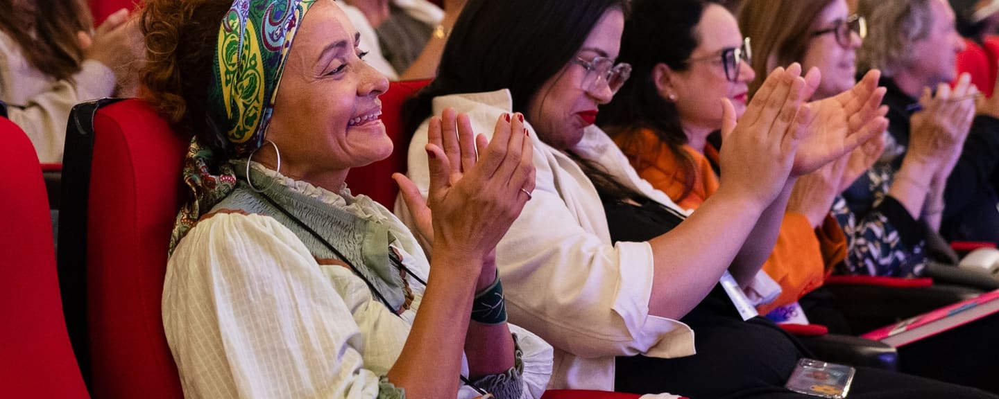 A woman in a printed headscarf smiles and claps in the audience at the 2nd Forum, with other participants applauding beside her