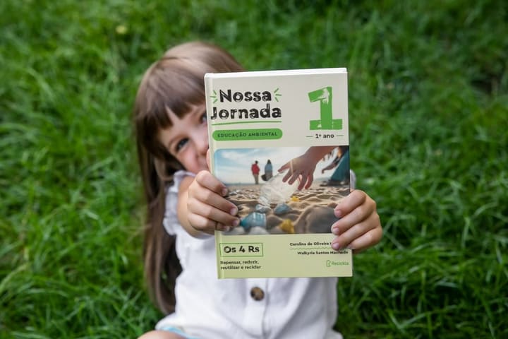 Criança segura o livro Nossa Jornada, do Recickla, num gramado.