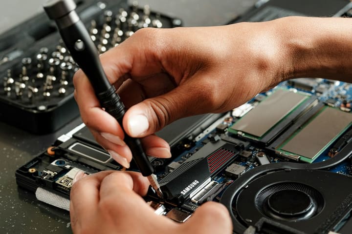 Mãos com chave de precisão desmontam um notebook aberto sobre a bancada, com o kit de pontas ao lado.