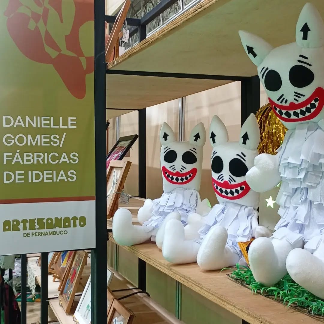 Bonecos brancos de tecido com sorriso vermelho numa prateleira, ao lado da placa Danielle Gomes / Fábricas de Ideias, Artesanato de Pernambuco