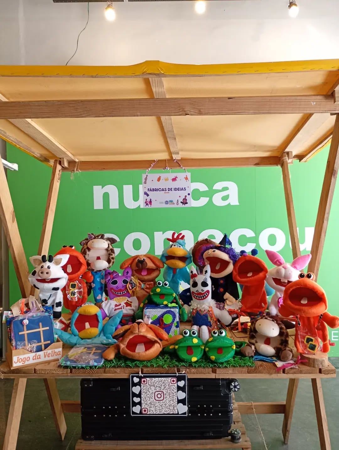 Bancada de madeira coberta de fantoches e bonecos de tecido coloridos, com a placa da Fábrica de Ideias pendurada no alto