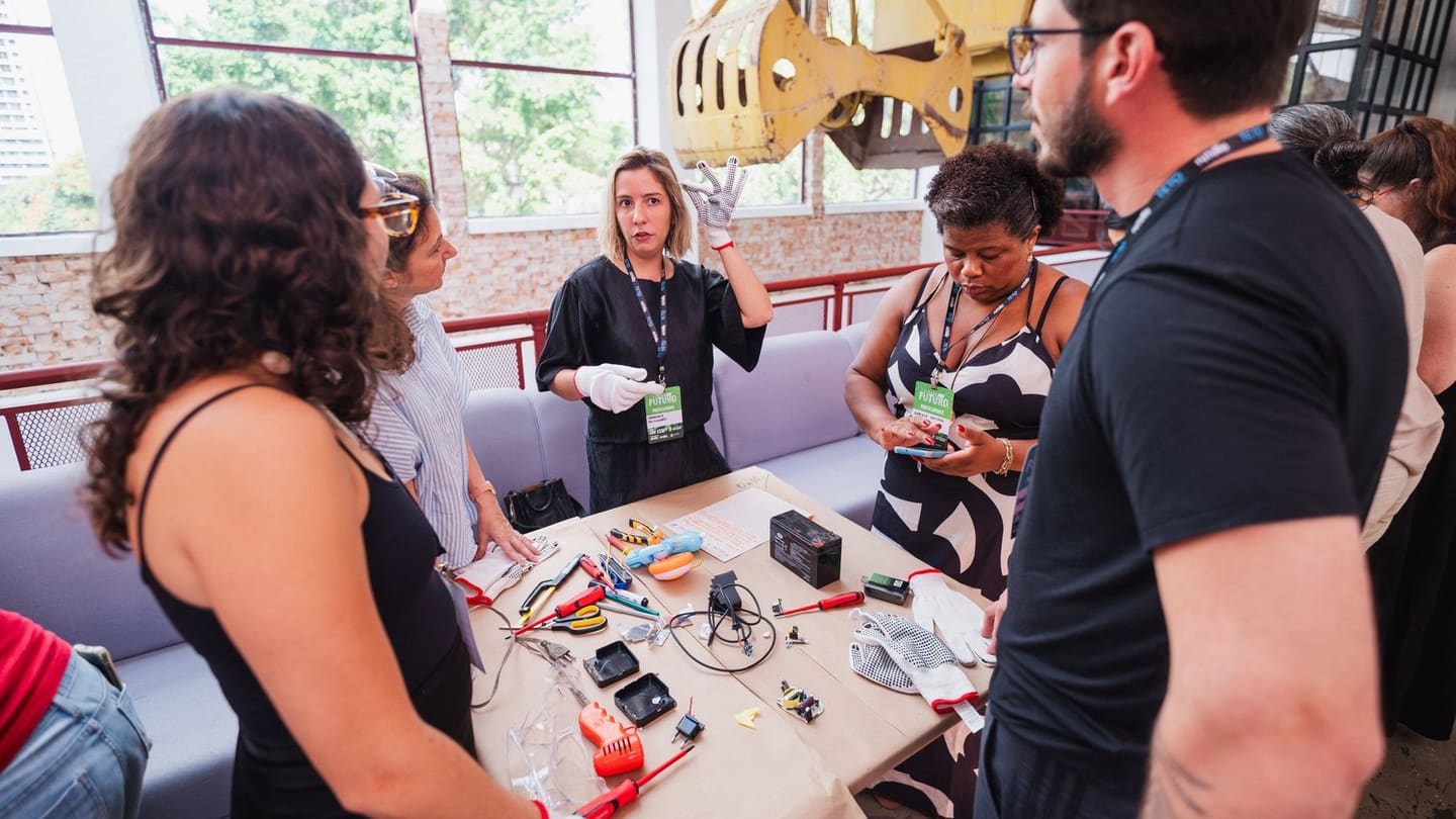 Facilitadora de luvas explica a desmontagem de eletrônicos a um grupo reunido em volta de uma mesa com ferramentas e peças.