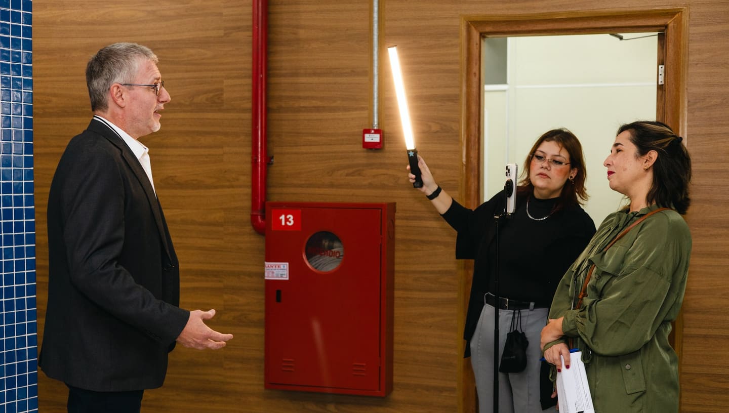 Bastidores da gravação da série no II Fórum: à esquerda, um entrevistado fala; à direita, uma integrante da equipe ilumina a cena com um bastão de luz e grava com o celular no tripé, ao lado de outra que acompanha com o roteiro na mão