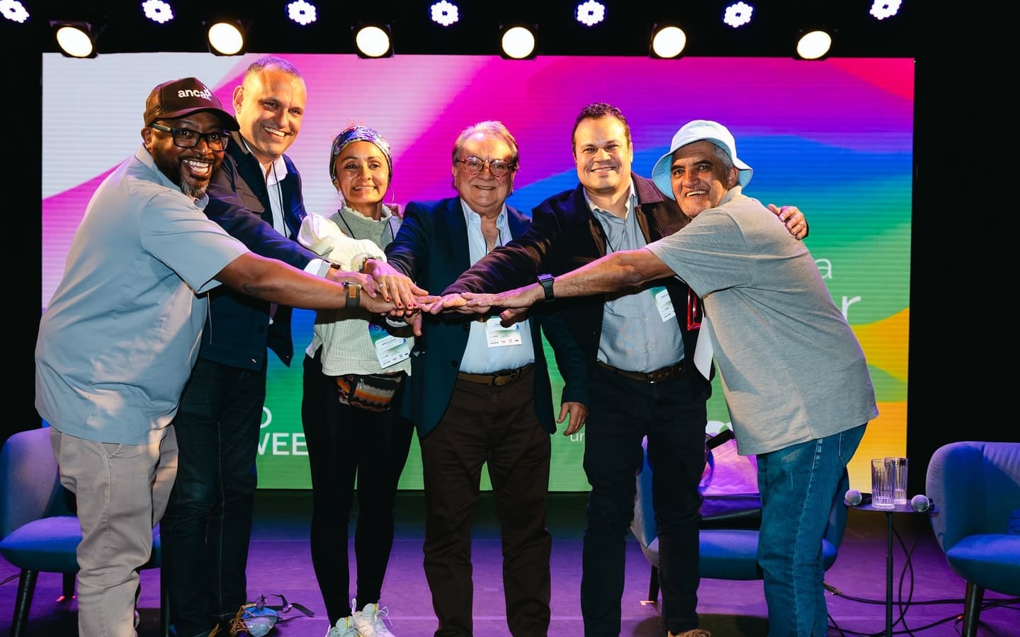 Six participants at the II Fórum stack their hands together on stage and smile at the camera.